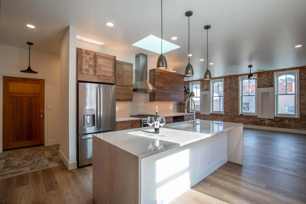 Modern kitchen by Durango Remodeling in Colorado featuring wood cabinetry, a large waterfall island, stainless steel appliances, pendant lighting, and exposed brick accent wall.