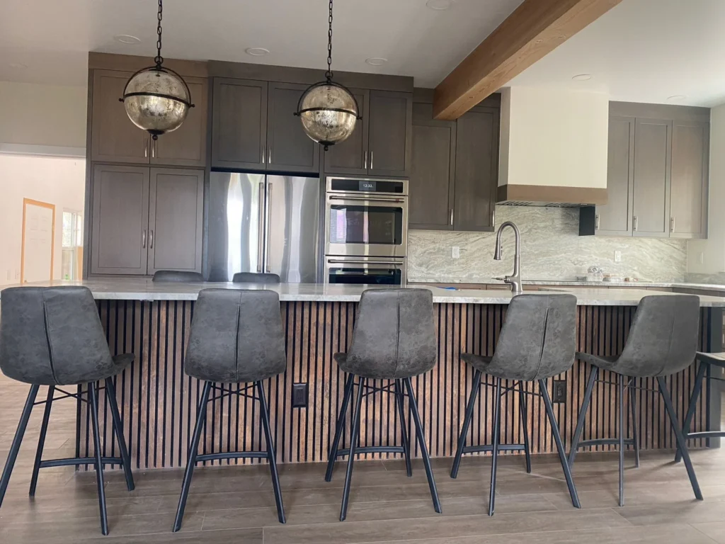 Modern kitchen by Durango Remodeling in Colorado with dark wood cabinetry, a long marble island with slatted wood detail and bar seating, stainless steel appliances, and globe pendant lighting.